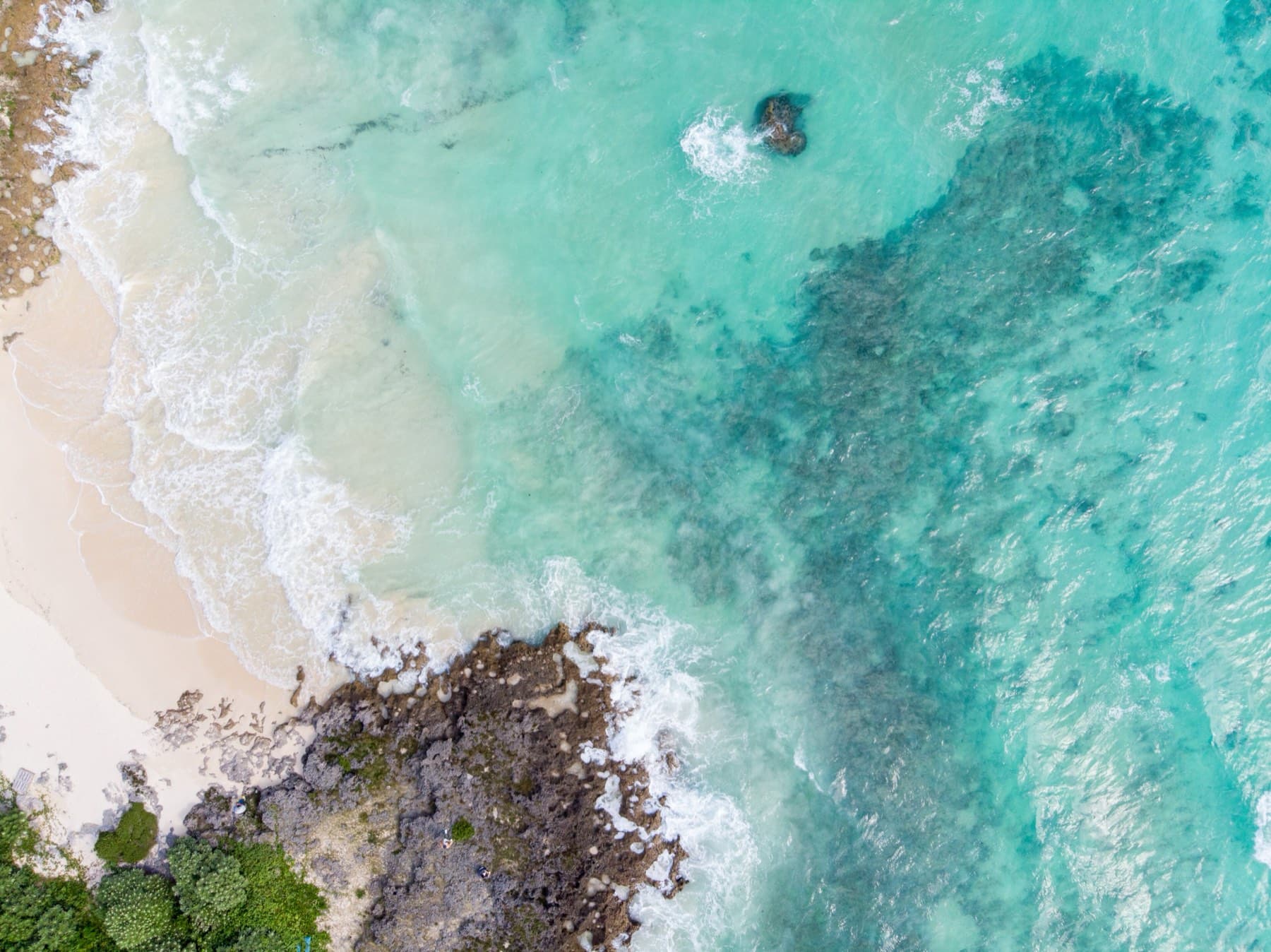 From above, white surf traces the edge of Toguchi no Hama where pale sand meets turquoise water and dark patches of reef.