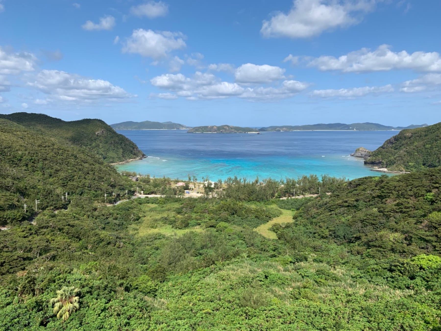 Forested hills open onto Tokashiku Bay on Tokashiki, and the shallow reef glows pale blue below a line of smaller islands on the horizon