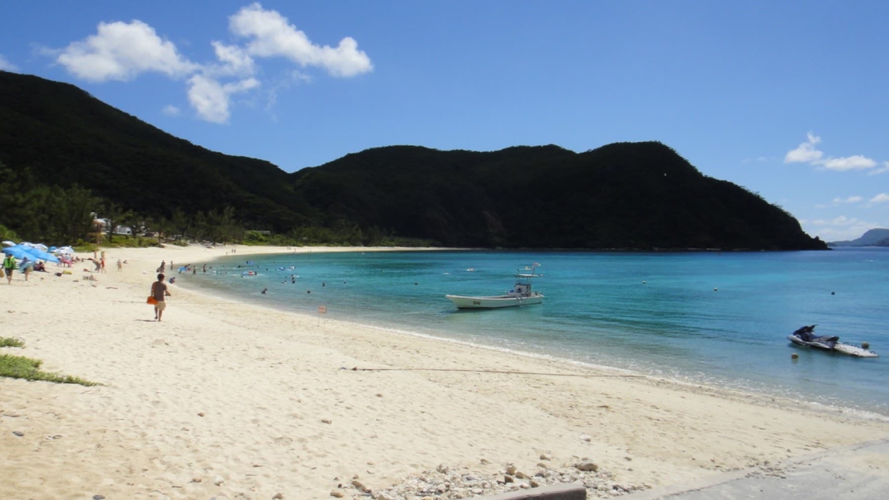 Small boats sit at anchor in the calm turquoise bay at Tokashiku Beach on Tokashiki as people walk the wide pale sand below dark green hills