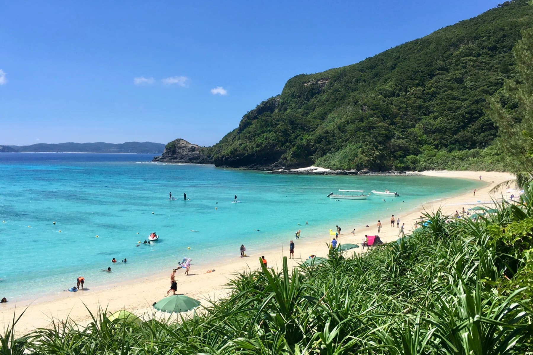Swimmers spread along the sand and shallows at Tokashiku Beach on Tokashiki, where a steep forested headland closes the north end of the bay
