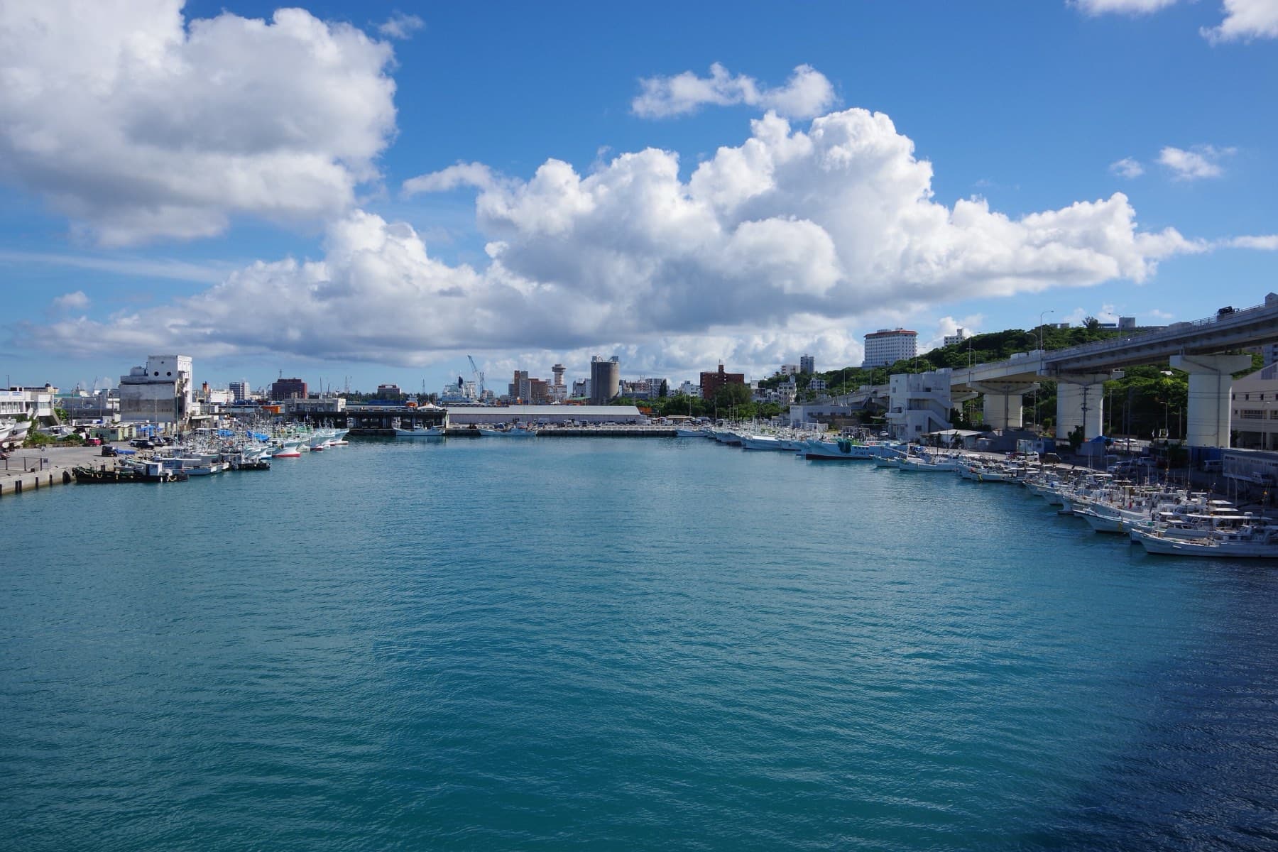 Fishing boats line both sides of the calm blue harbour at Tomari under big summer clouds, and the city of Naha rises beyond the water