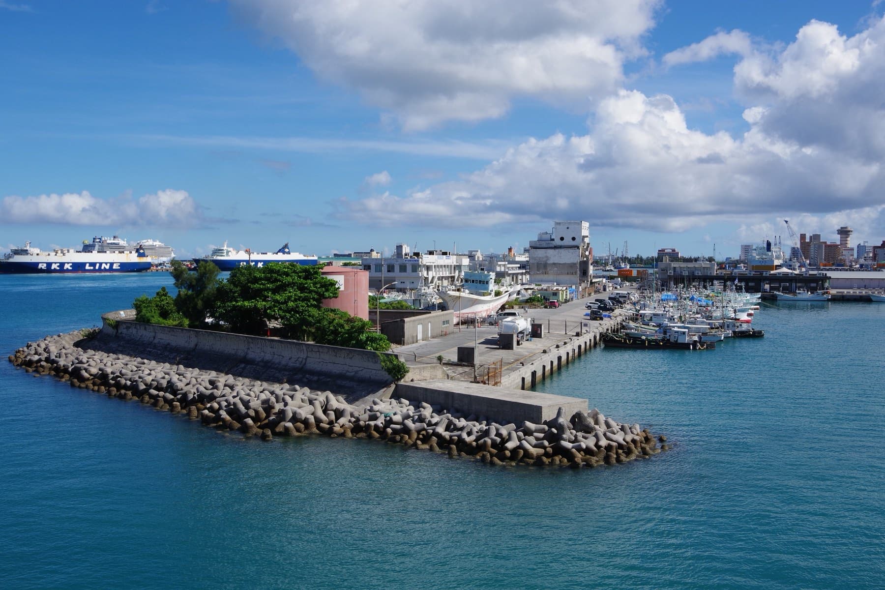 A rock breakwater curls into the blue water of Tomari harbour while large ferries wait at the quays behind under a bright sky