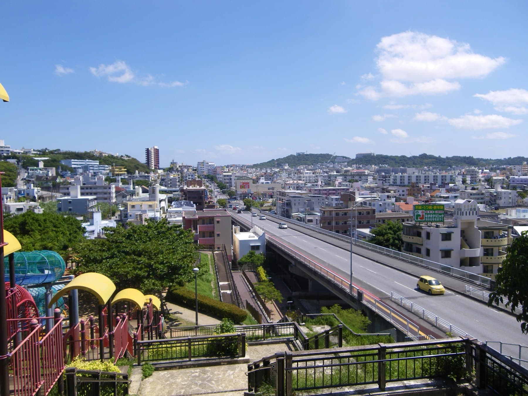 From the Navy Headquarters Park hill the dense white houses of Tomigusuku and an elevated road stretch toward the hills under a blue sky