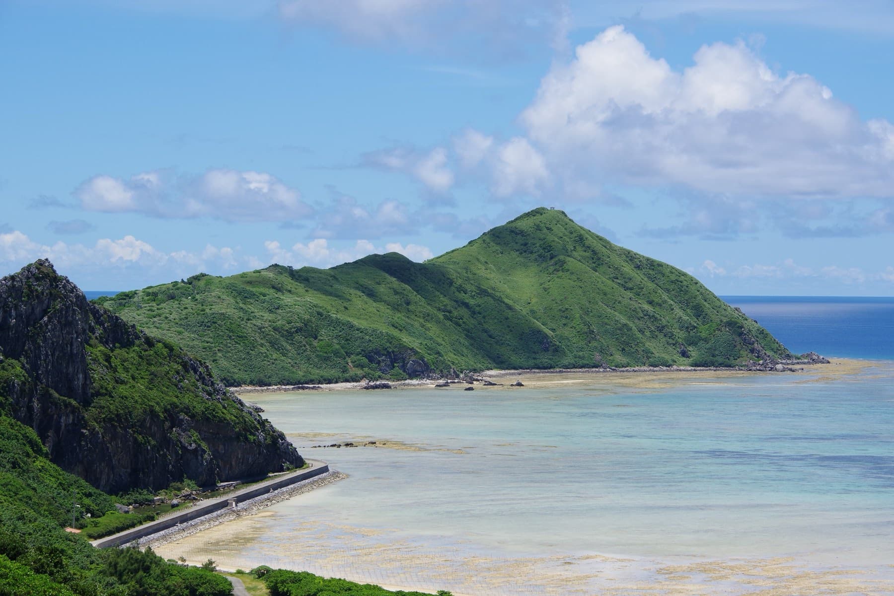 The green dome of Nishimui rises over a wide shallow bay on Tonaki, and a coast road runs along the sea wall below a dark cliff