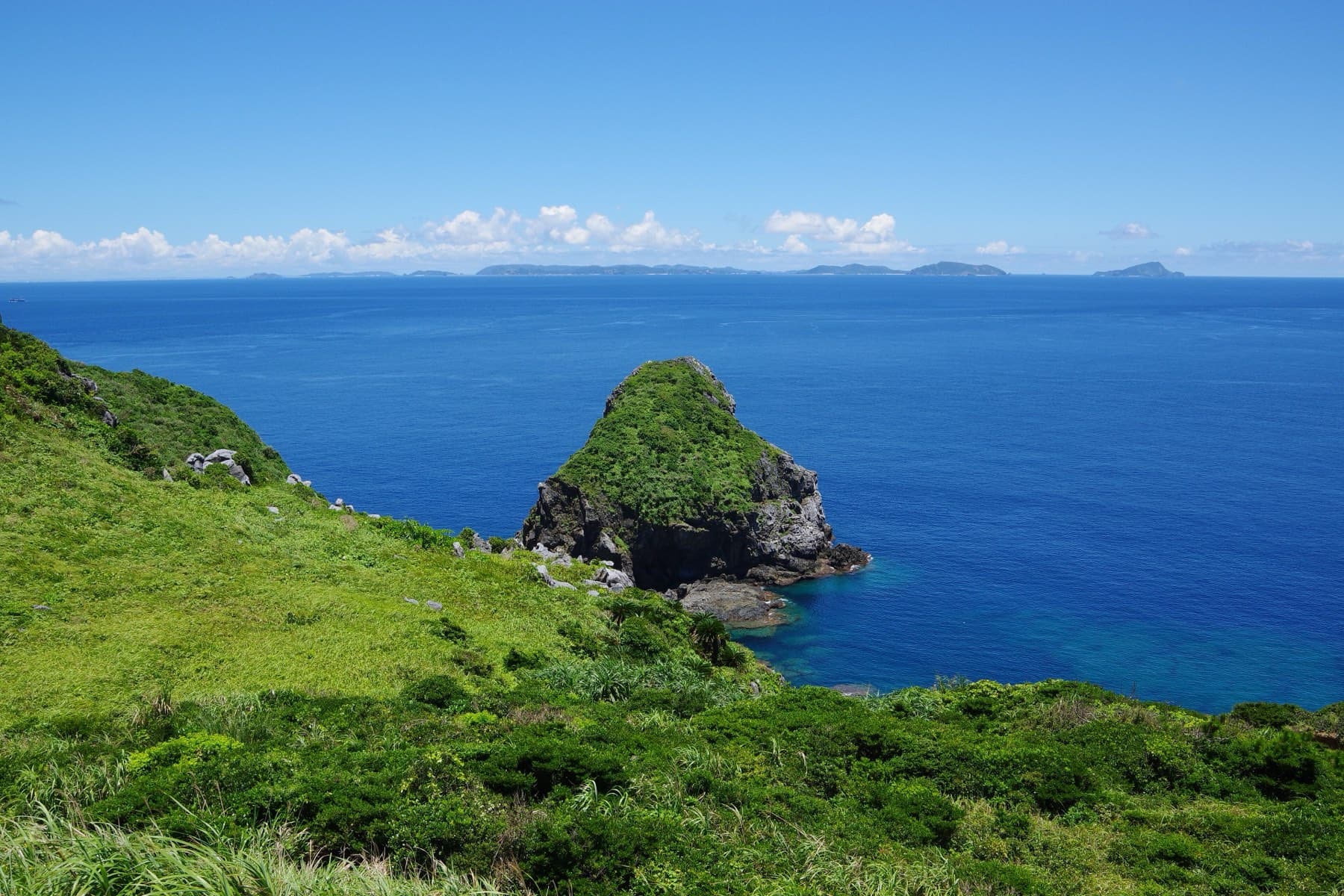 Grassy slopes fall to a rocky islet at Shimajirizaki on Tonaki, and the low line of the Kerama Islands sits on the horizon across deep blue water