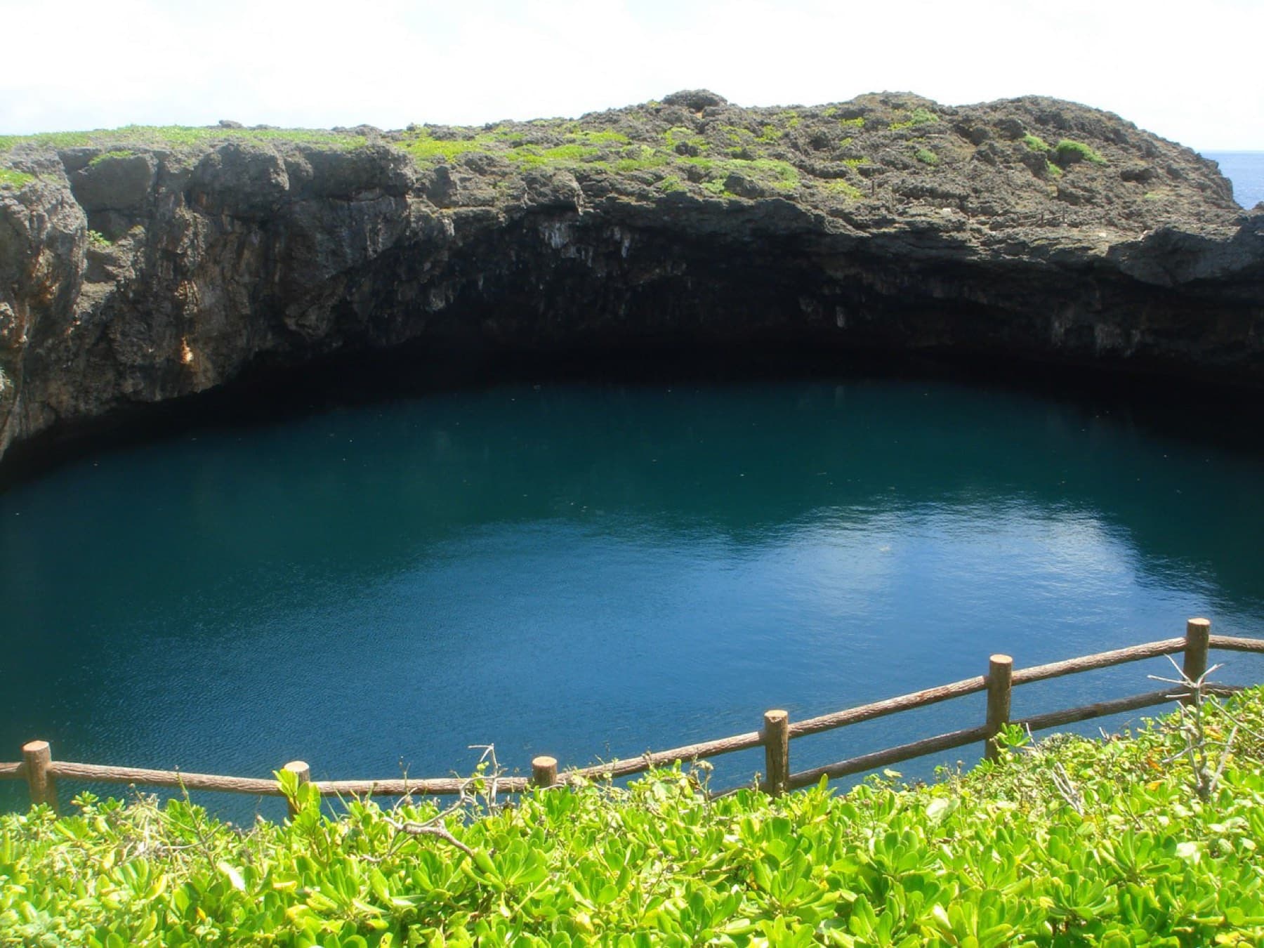A wooden fence runs along the rim above the deep blue water of a Toriike pool beneath an overhang of rough limestone.