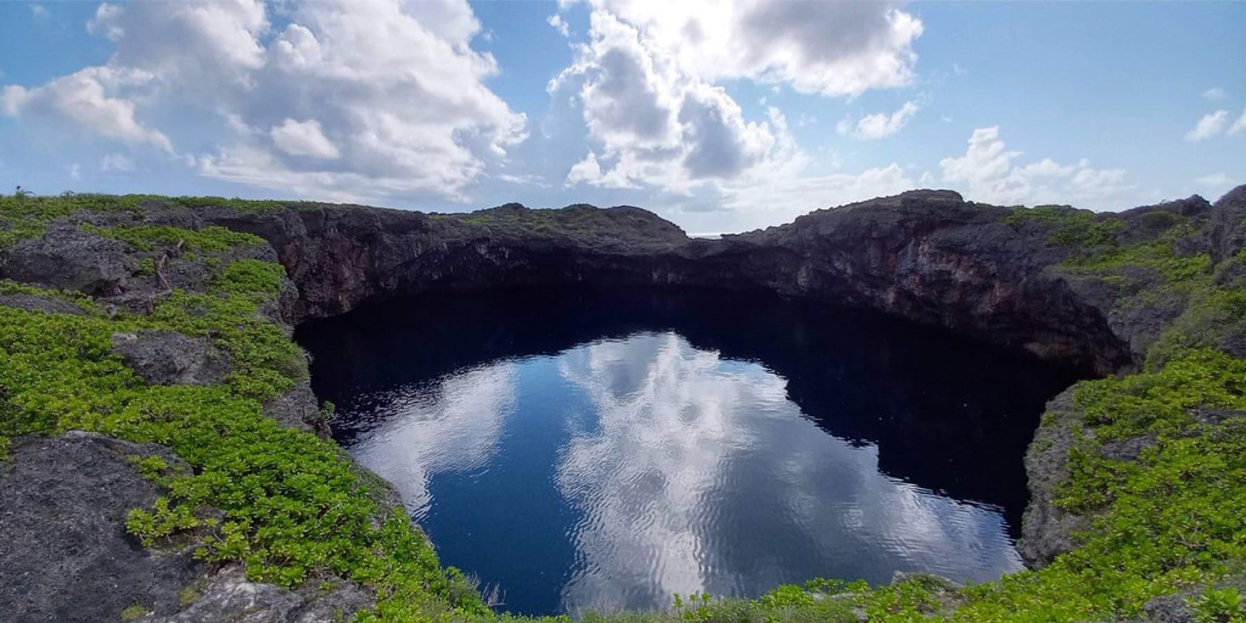 Low cliffs of grey limestone ring one of the Toriike sinkholes on Shimoji, and white clouds reflect in its dark, still water.