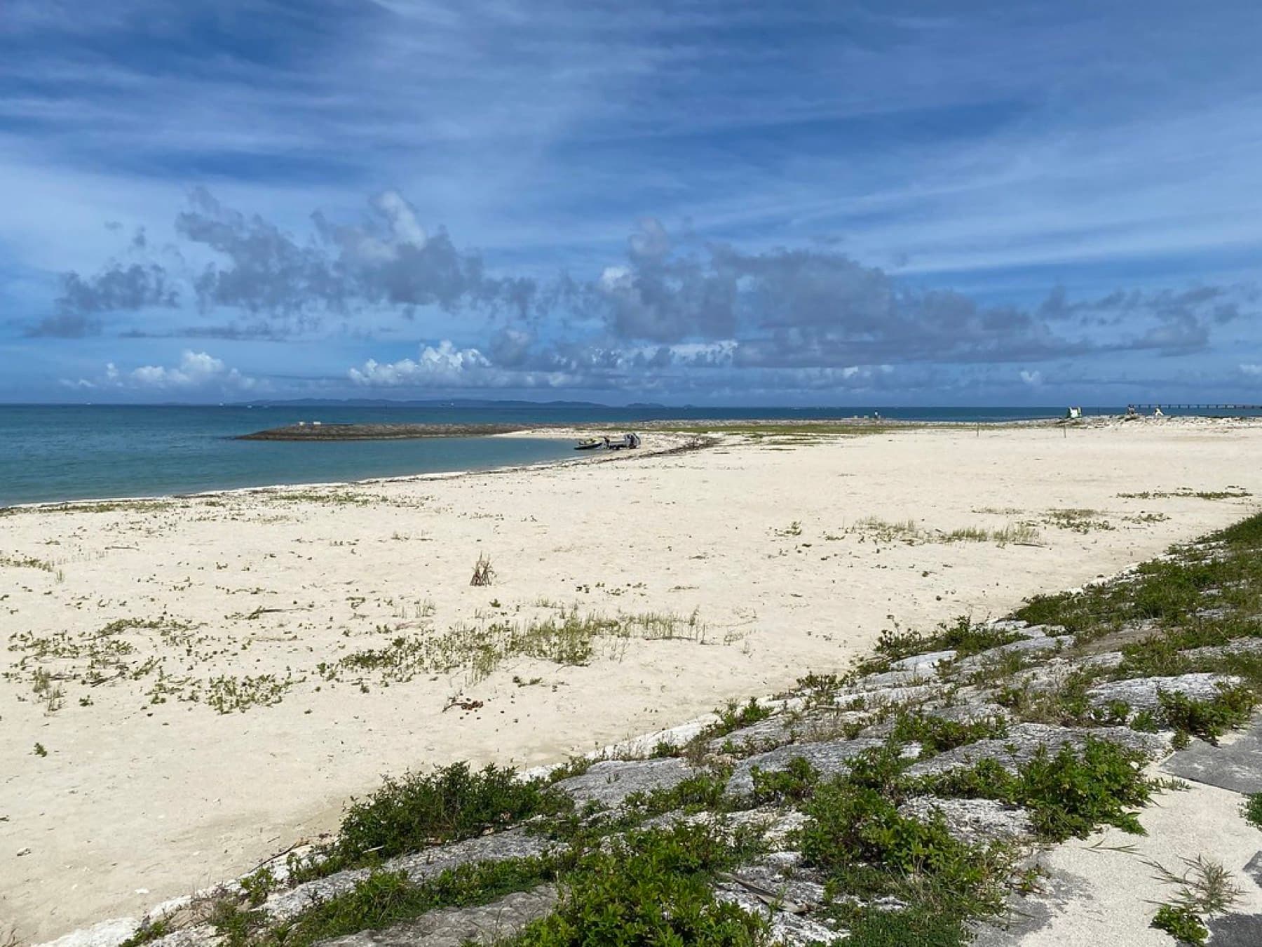 Pale sand stretches along Toyosaki Churasun Beach in Tomigusuku toward a low breakwater, with green dune plants in front and broken cloud over the sea