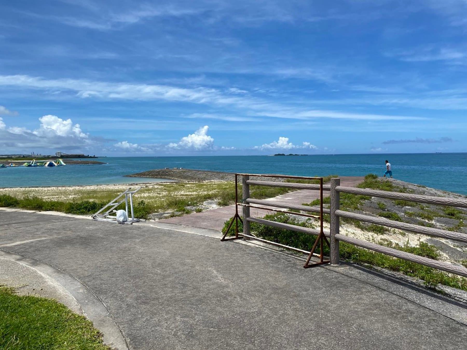 A paved seaside path with a wooden railing curves along the Toyosaki shore in Tomigusuku above blue water and a sandy beach under bright summer sky
