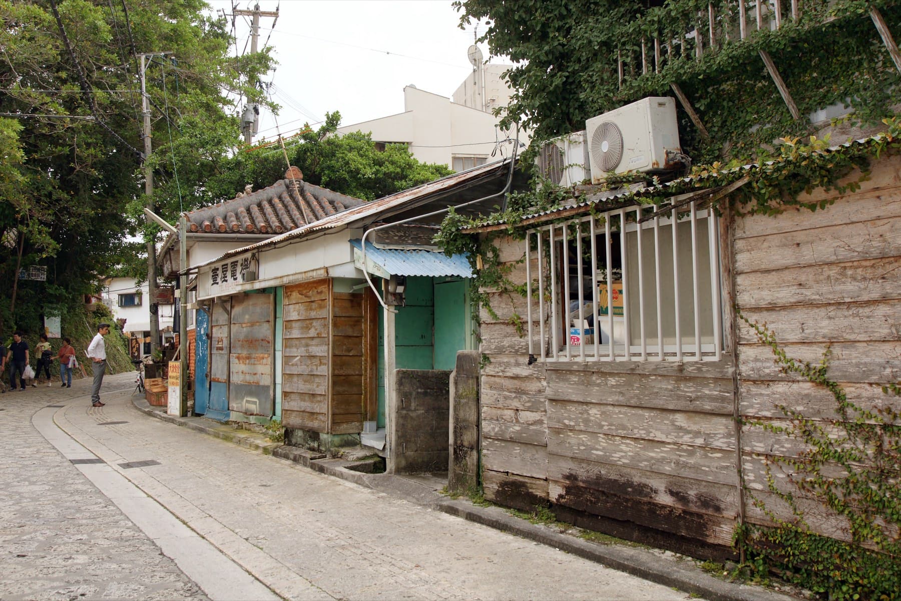 Weathered wooden houses and a small tiled roof sit along a quiet paved lane in Tsuboya, and a few people walk up the street ahead