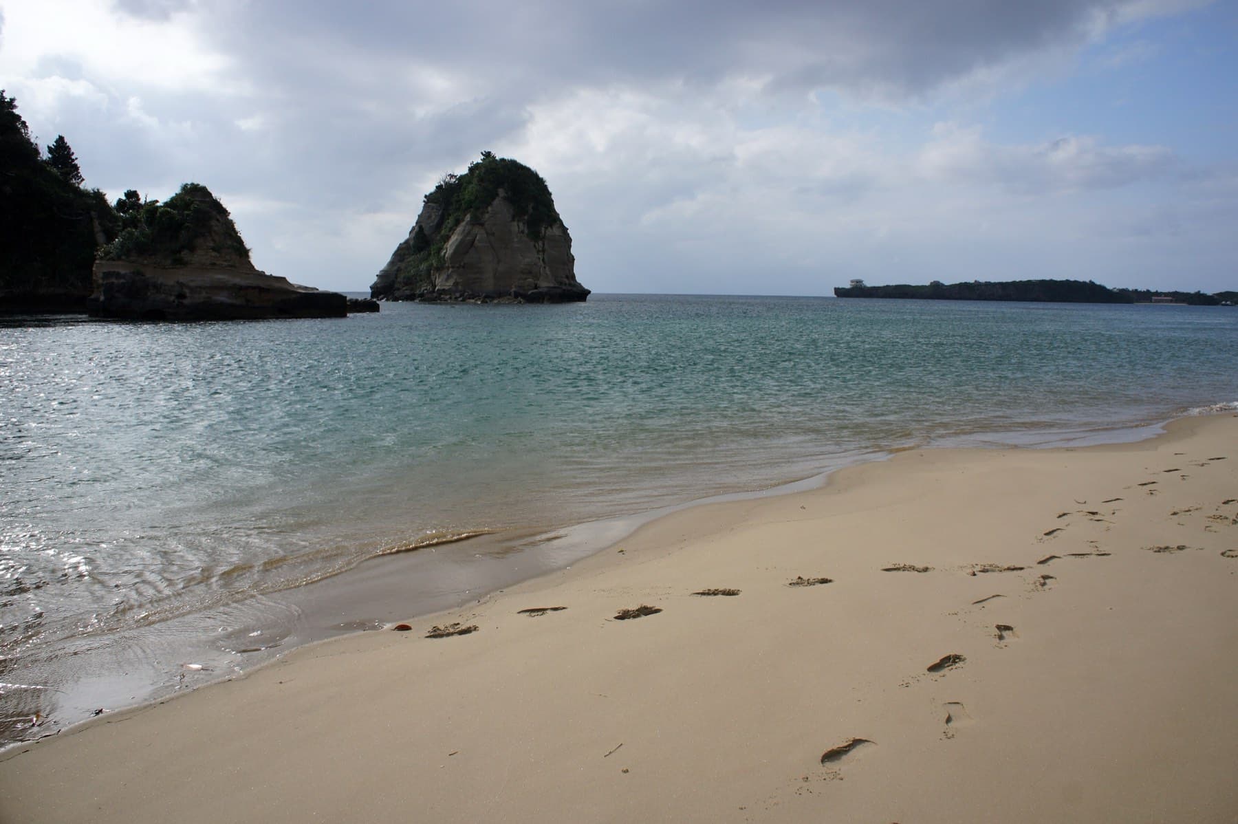 Two rock stacks crowned with trees rise from the calm water at the end of Tsukigahama, where footprints mark the soft sand.