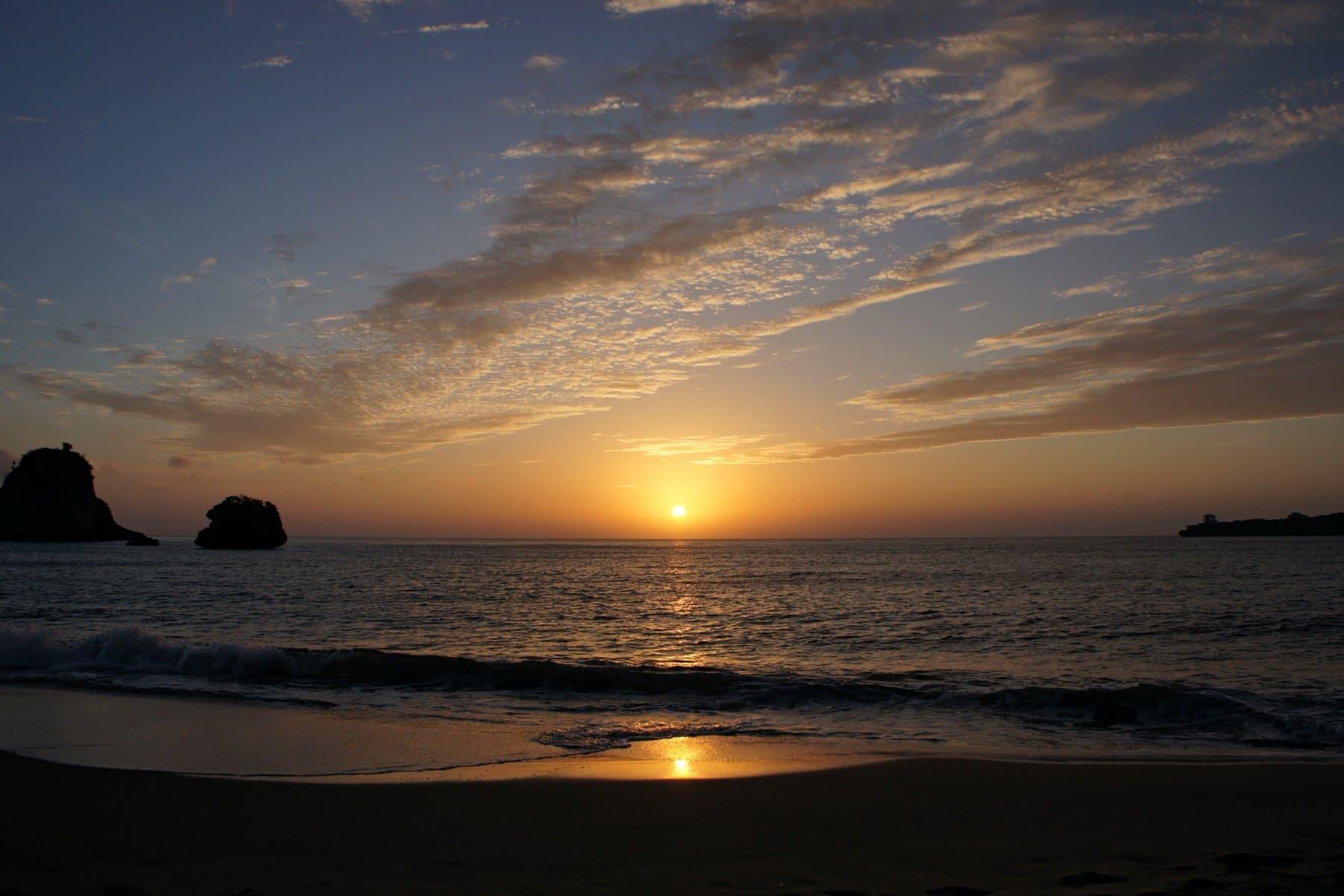 The sun sets between scattered cloud over Tsukigahama on the north coast of Iriomote as dark rock stacks stand in the bay.