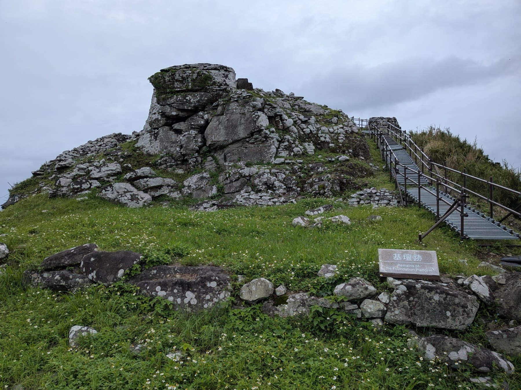 A stack of weathered stones marks the summit of the Uegusuku Castle ruins on Kume-jima, where a metal stairway climbs past grass and scattered boulders