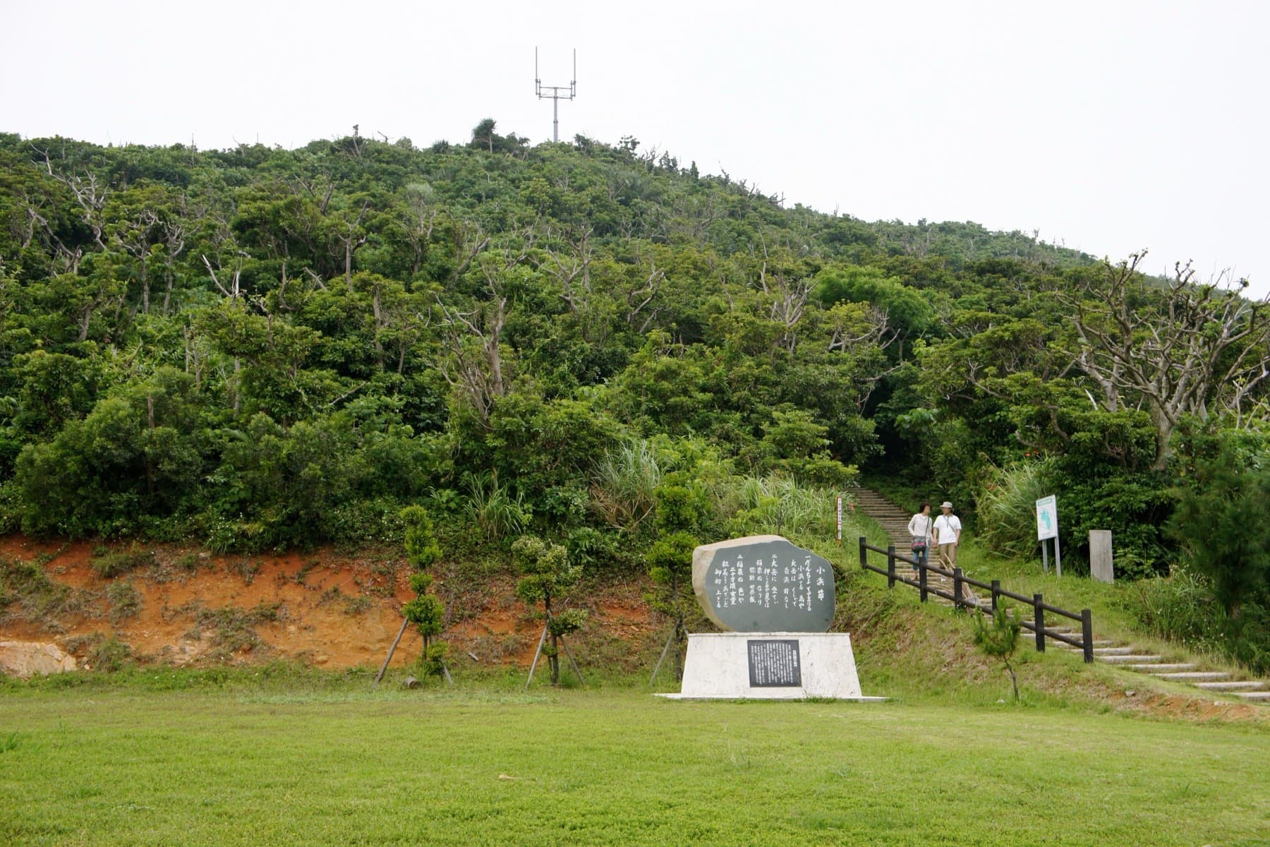 Two walkers pass a stone monument where the railed path starts up the wooded slope of Ufudaki in the middle of Kohama.