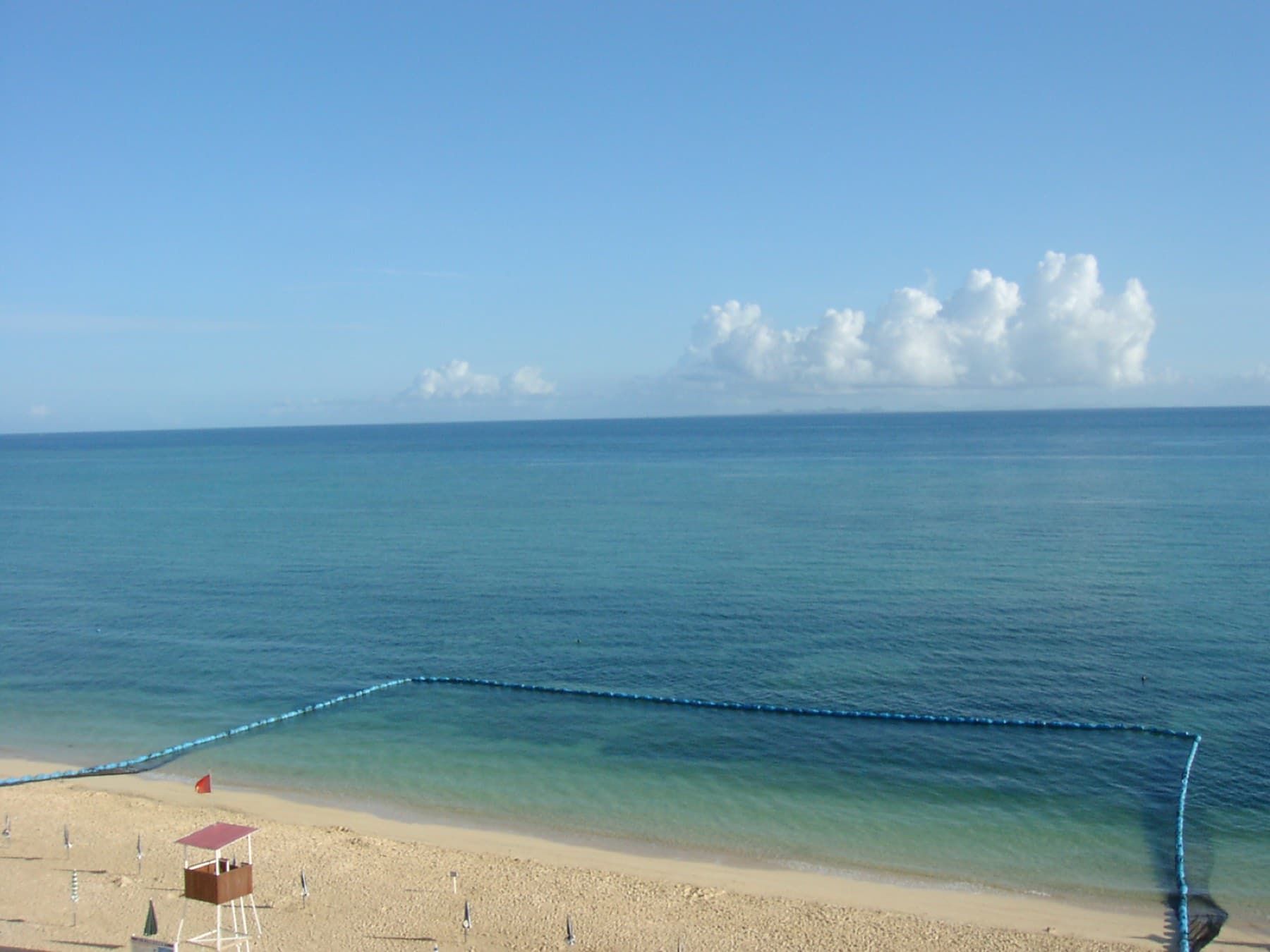 A jellyfish net marks a calm swimming area off Uppama Beach in Nakijin on a summer morning, with a lifeguard tower on the pale sand