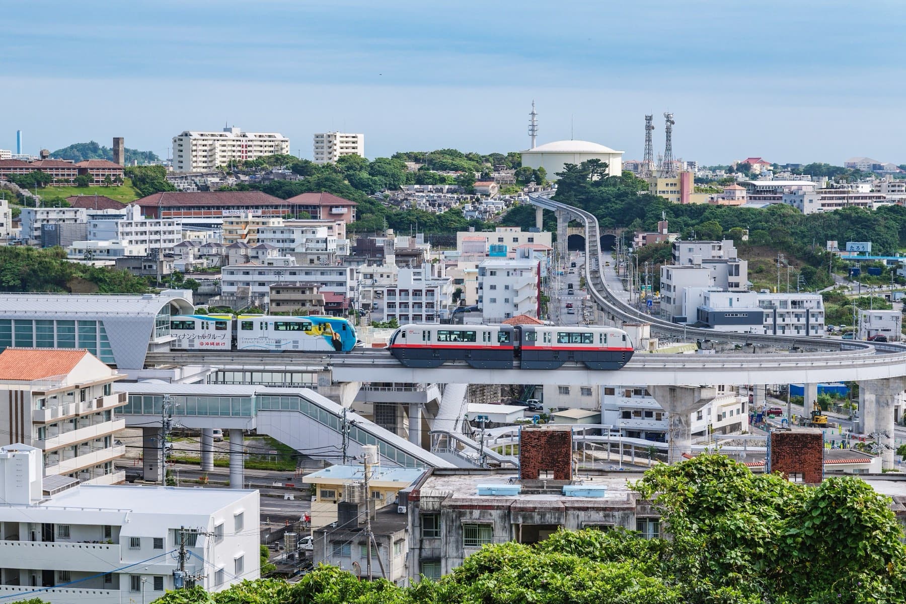 Two monorail trains pass at Urasoe-Maeda station while the guideway climbs away through the hillside houses and green slopes of Urasoe