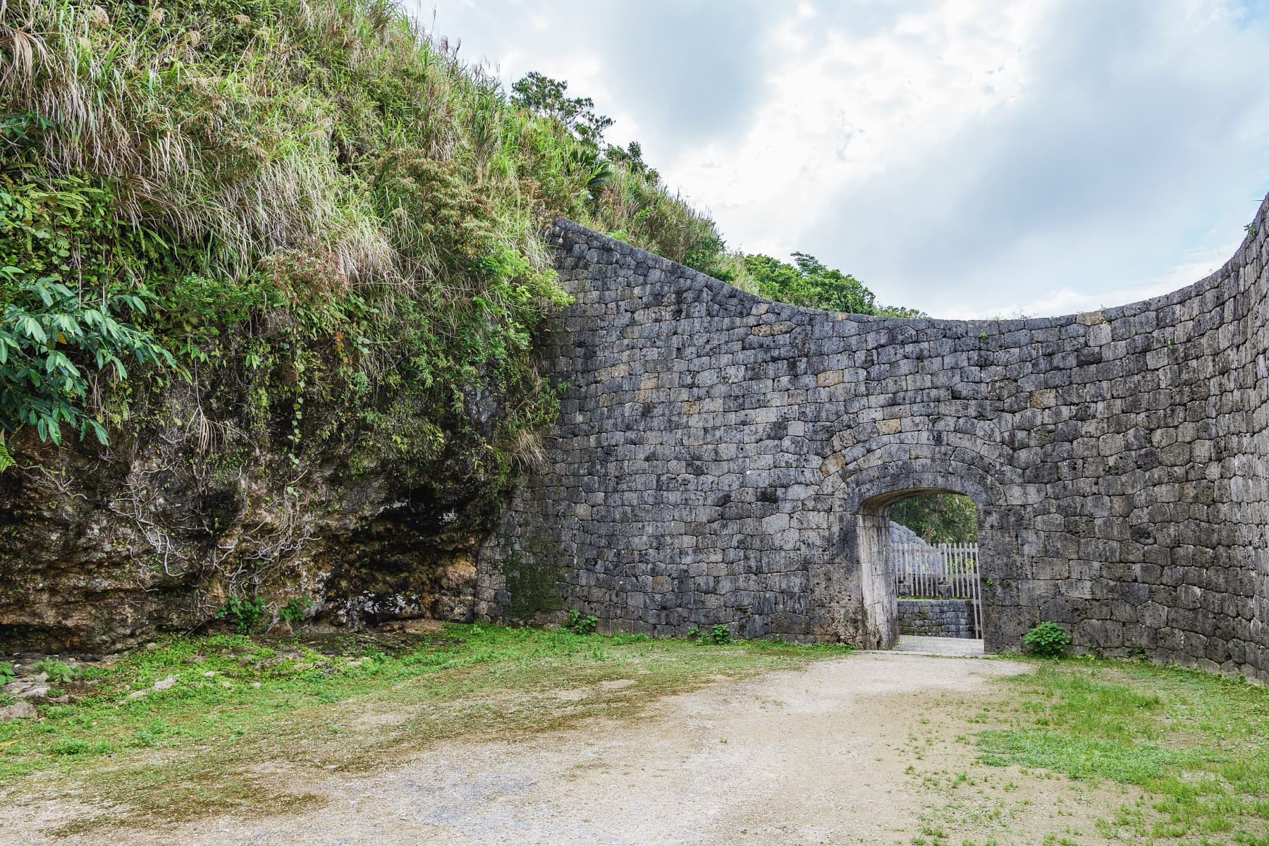 A curved wall of fitted stone with a small arched gateway closes the forecourt of the Urasoe Yodore tombs at the foot of a grassy cliff