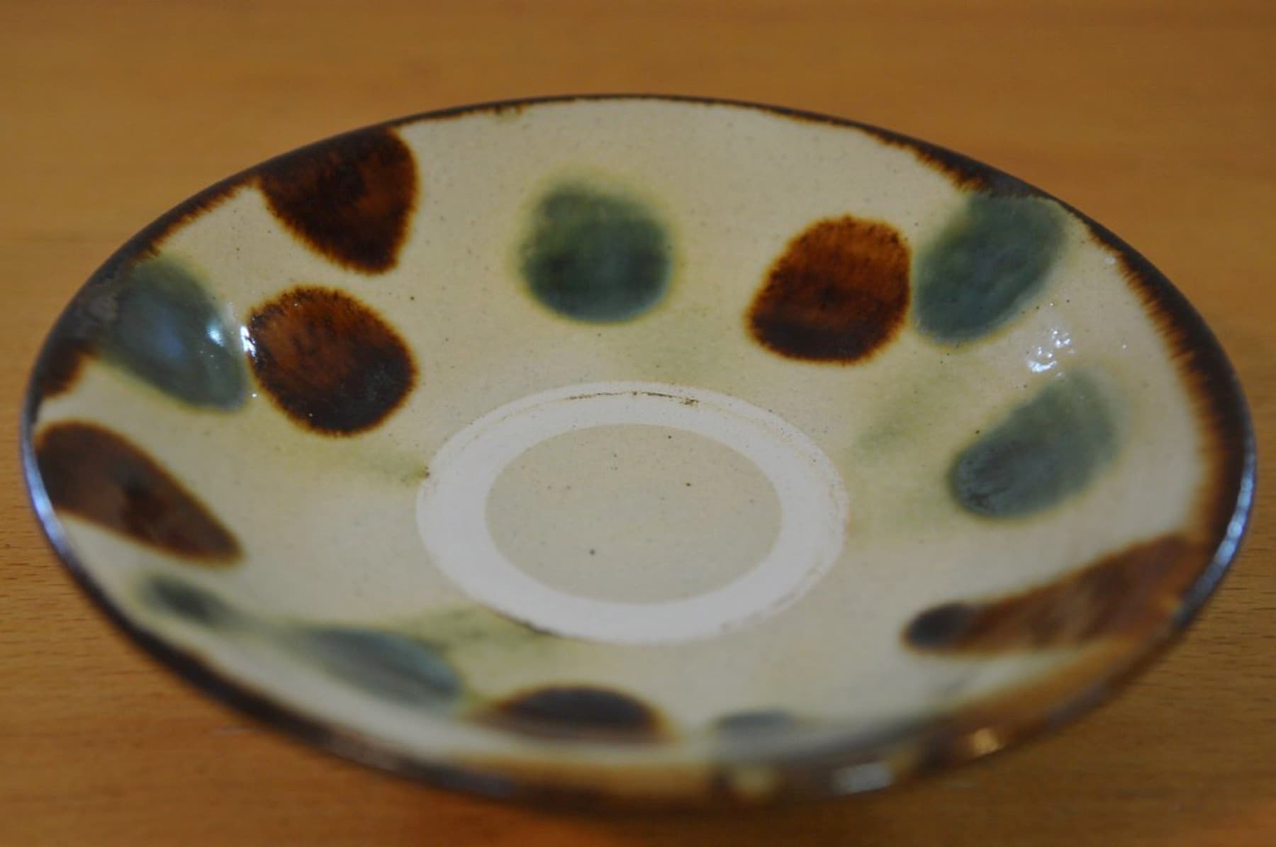 A shallow stoneware dish from the Kitagama kiln in Yomitan carries alternating brown and green glaze dots around a cream glaze with an unglazed ring at the centre