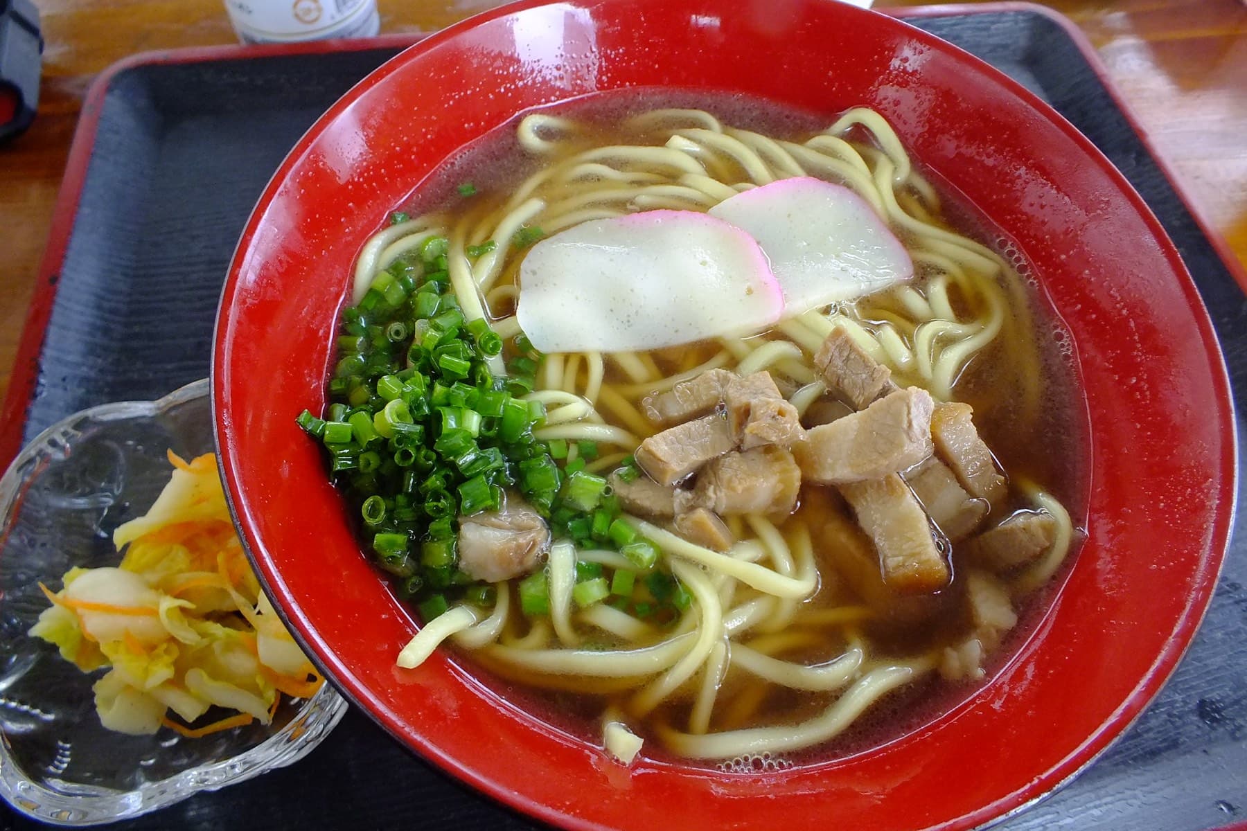 A red bowl of Yaeyama soba on Ishigaki holds round noodles, diced pork, pink fish cake and a heap of green onion beside a dish of pickles