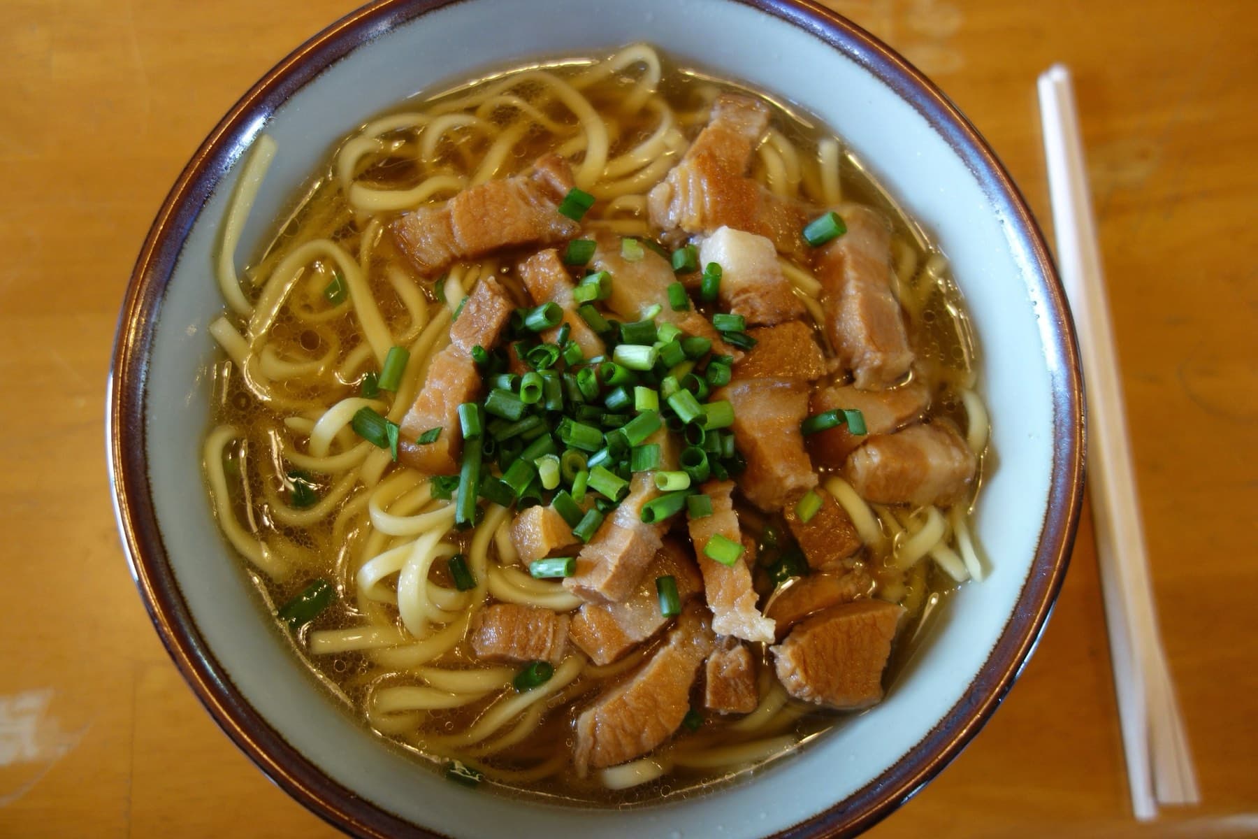 Thin strips of simmered pork and chopped green onion cover round wheat noodles in a light brown broth, with chopsticks laid beside the bowl
