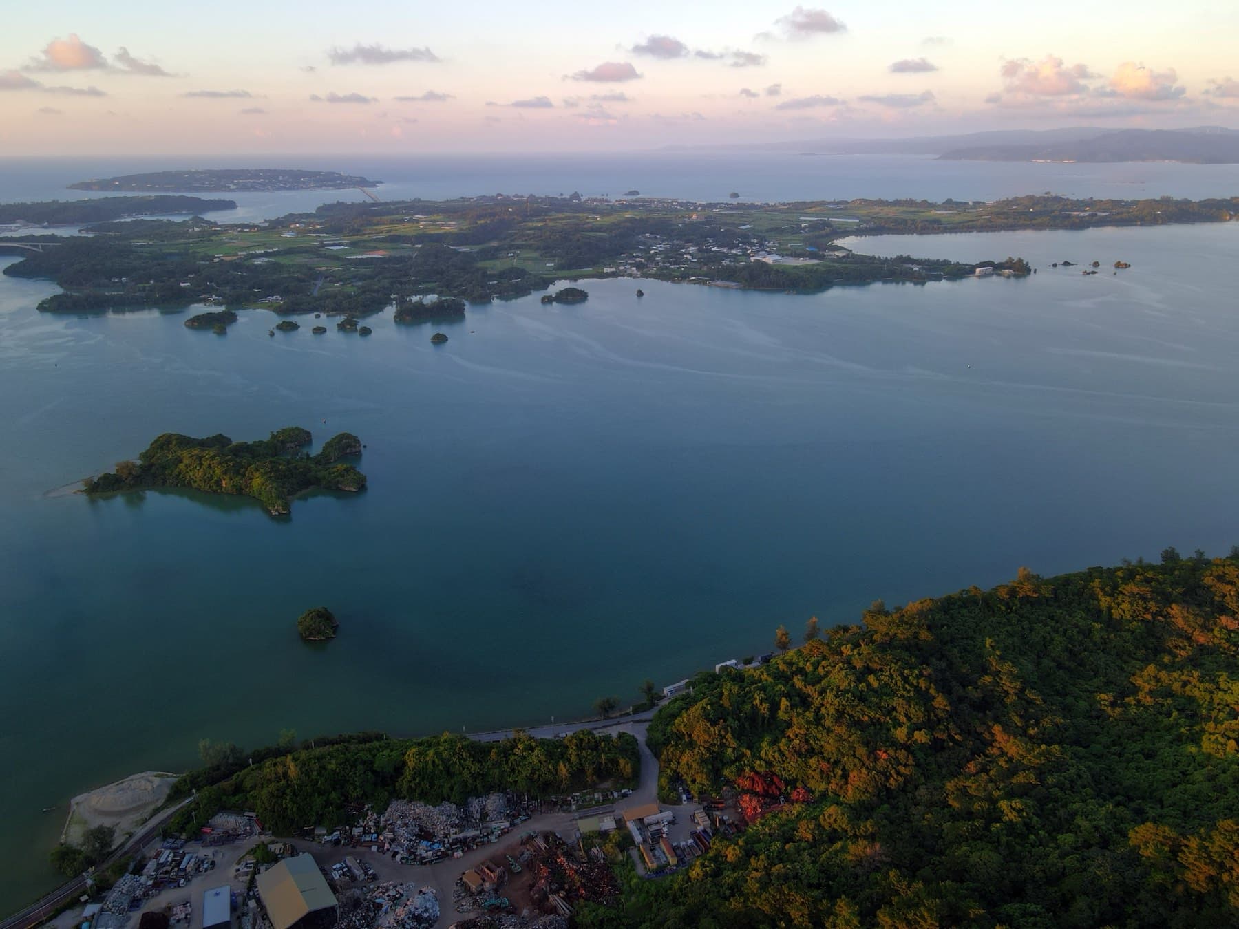 At dusk the calm Haneji Inland Sea spreads below a drone, with small wooded islets in the water and the flat farmland of Yagaji beyond under pink cloud