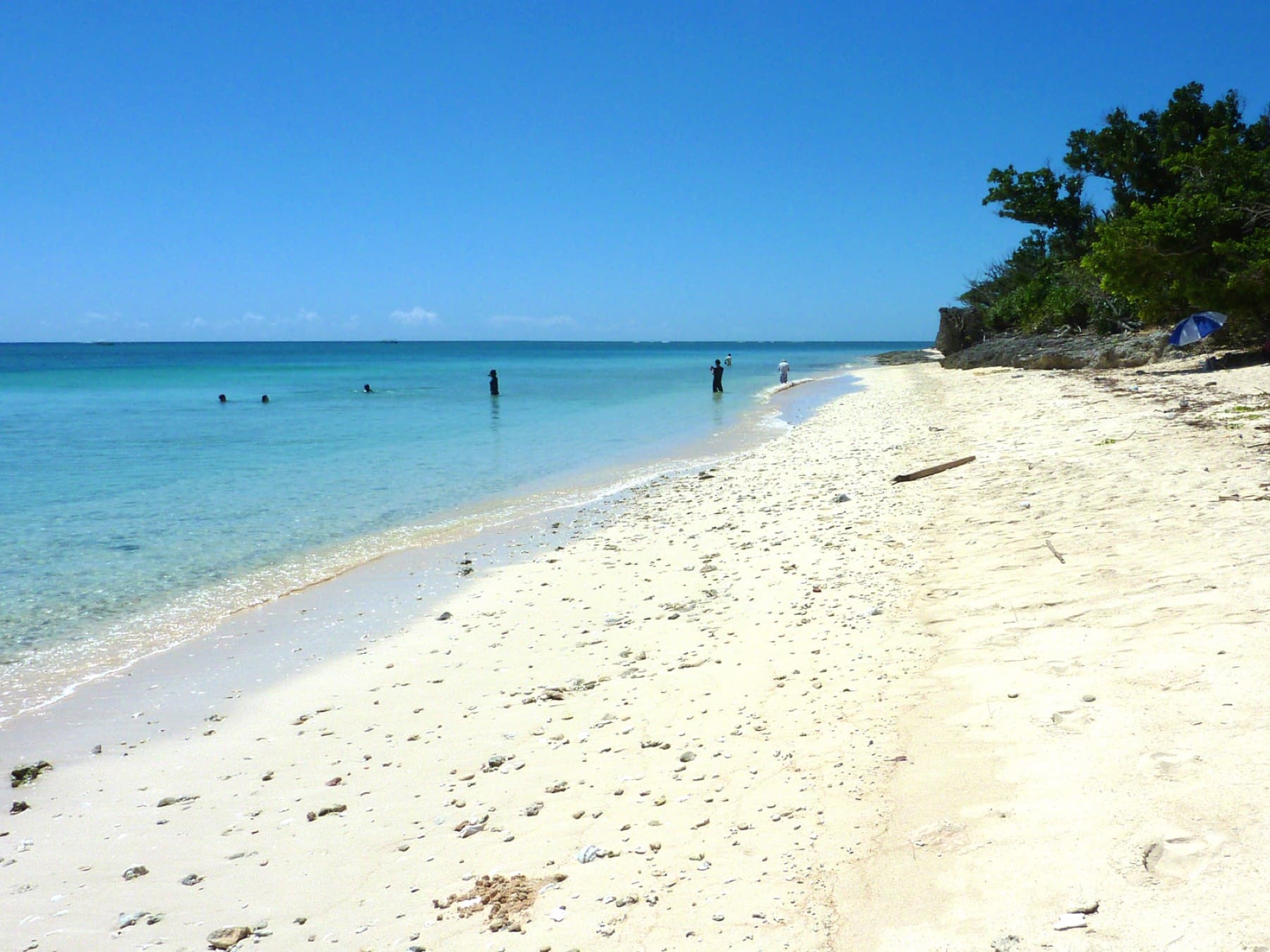 A strip of bright coral sand runs beside clear turquoise water at Yara-hama on Hatoma as a few people wade near the far end.