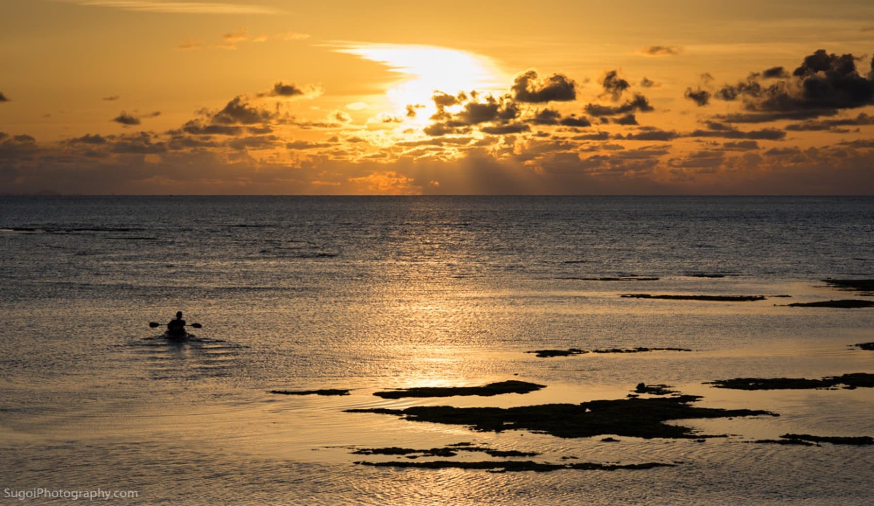 A lone paddler crosses golden water at sunset off Toguchi in Yomitan, where dark reef rocks break the calm surface below a sky of glowing cloud