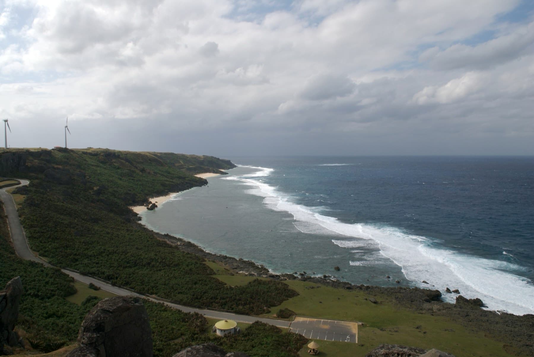 Lines of white surf break along the reef of a long curving bay below the grassy hills of Yonaguni under a heavy sky.