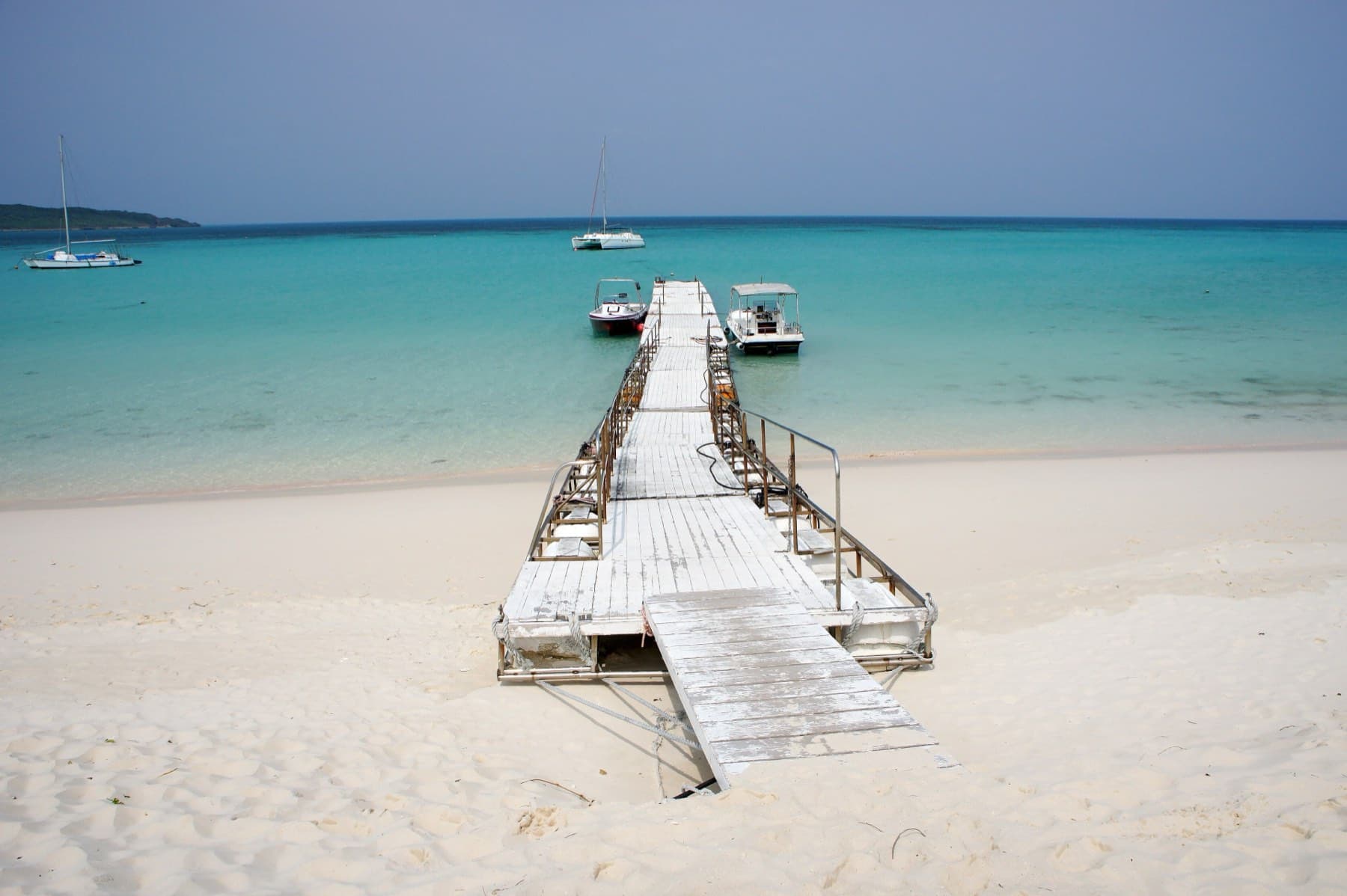 A weathered boardwalk leads from the white sand of Yonaha Maehama out to small boats moored over pale blue shallows.