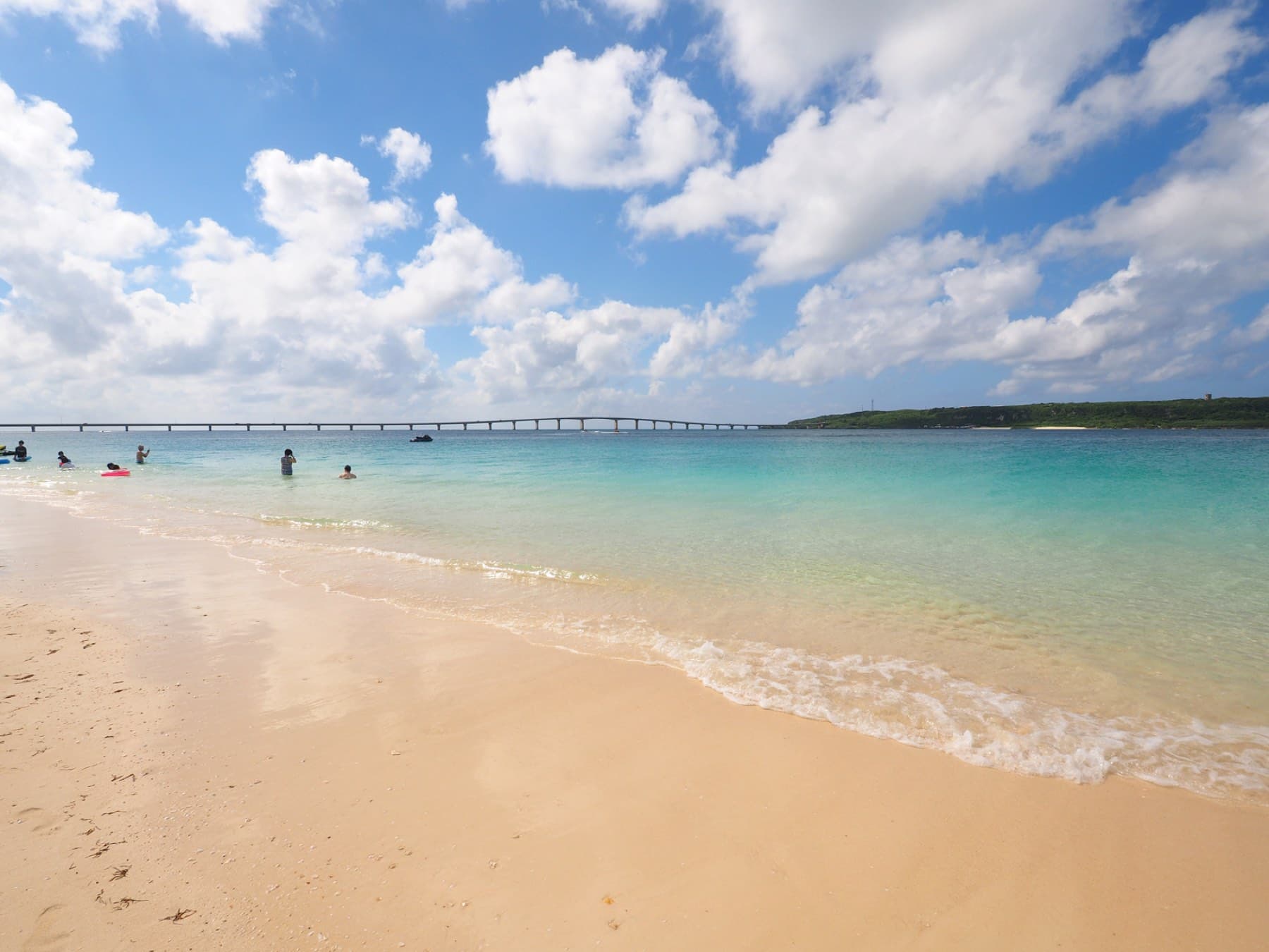 Gentle surf rolls onto the soft sand of Yonaha Maehama under bright summer cloud while the long Kurima Bridge crosses the water behind the swimmers.