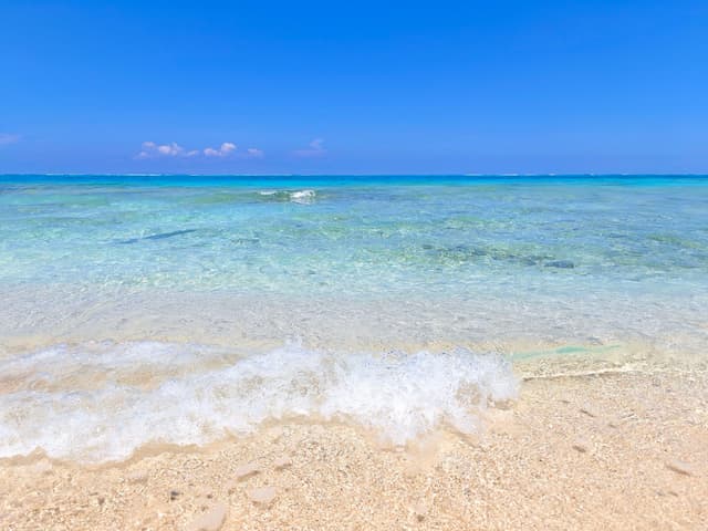 A gentle wave spreads foam over white sand on Yoron as the shallow water runs clear and pale toward the reef edge.