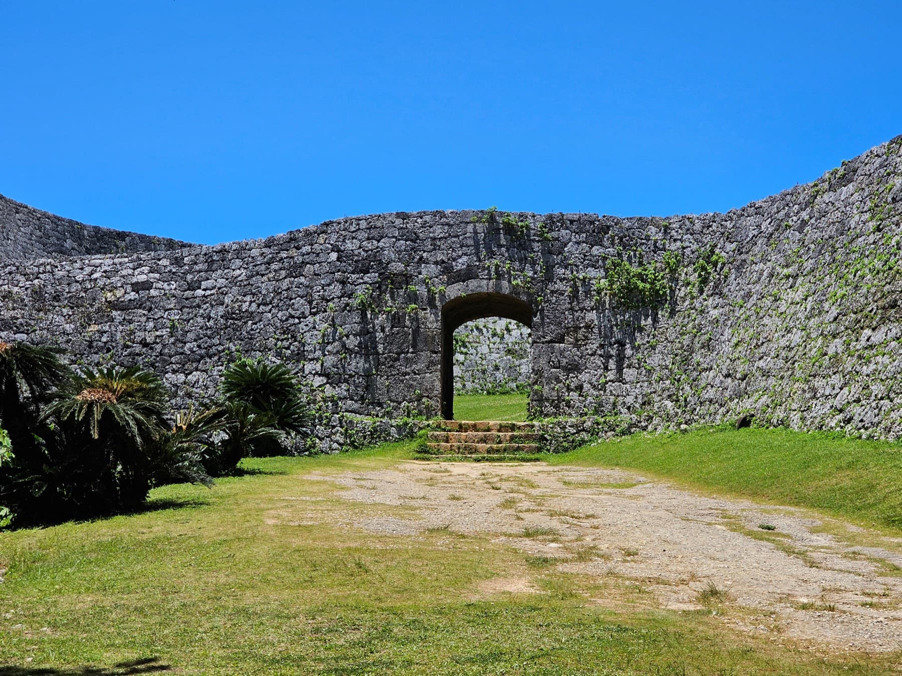 A low arched gate pierces the curving limestone wall of Zakimi Castle in Yomitan, with cycads beside it and a sandy path across the lawn under a deep blue sky