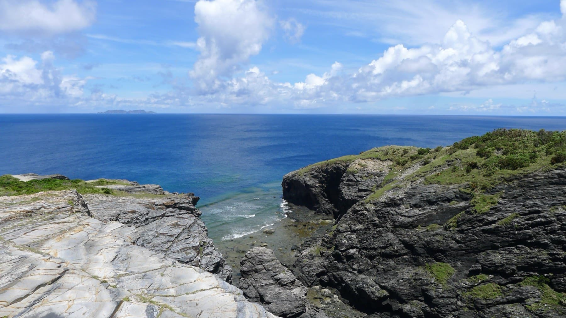 Layered grey rock forms a cliff edge on Zamami, and the open sea stretches deep blue to a low island on the far horizon