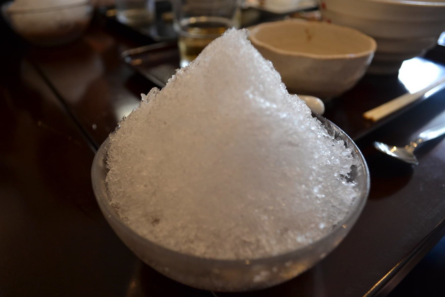A tall cone of finely shaved ice for Okinawa zenzai fills a frosted glass bowl on a dark polished table in soft window light